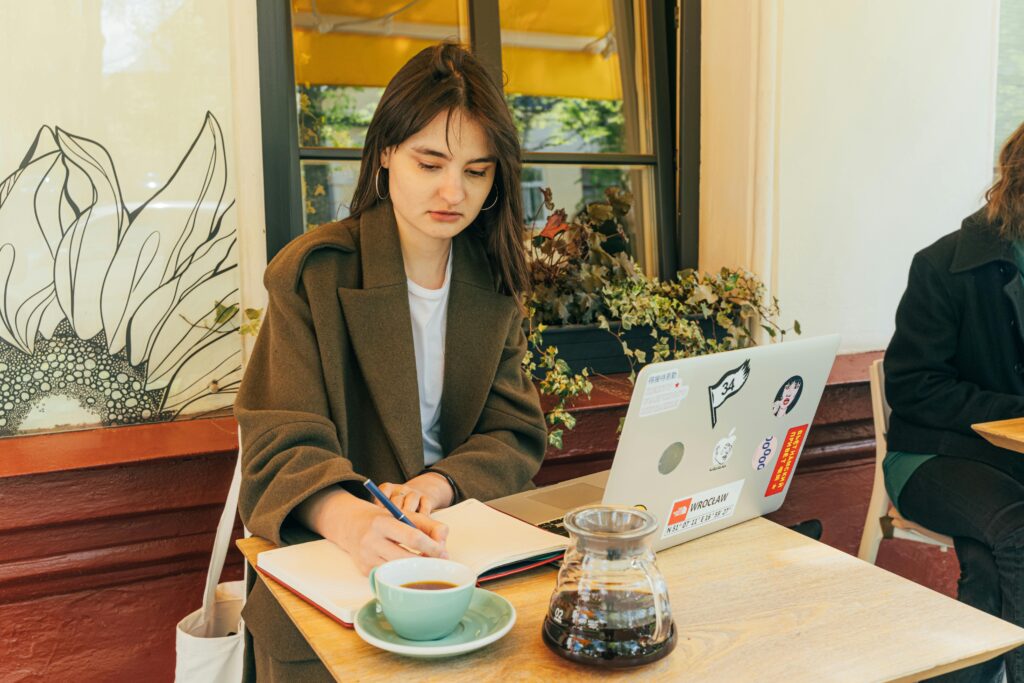 Young woman working at café, writing notes beside laptop with coffee cup and pot on table.