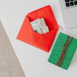 A green gift box and red envelope with cash on a desk beside a laptop, emphasizing holiday or financial themes.